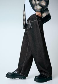 Széles szárú fekete farmernadrág kontrasztos fehér varrással, vastag fekete cipőkkel párosítva. A nadrág öt zsebbel és fémes díszítéssel rendelkezik.