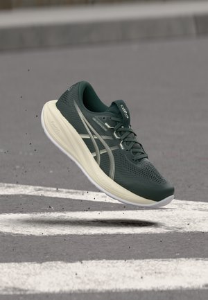 Chaussure de course vert foncé avec une semelle blanche en l'air au-dessus d'une route asphaltée avec des lignes peintes blanches, montrant la semelle et les bandes latérales de la marque.