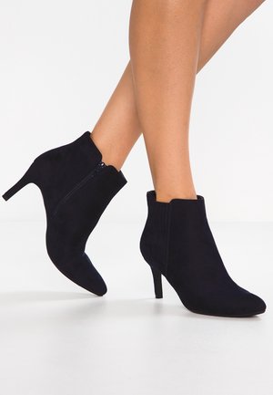 Woman wearing black suede ankle boots with side zipper and slim mid-height heels, standing on a white floor.