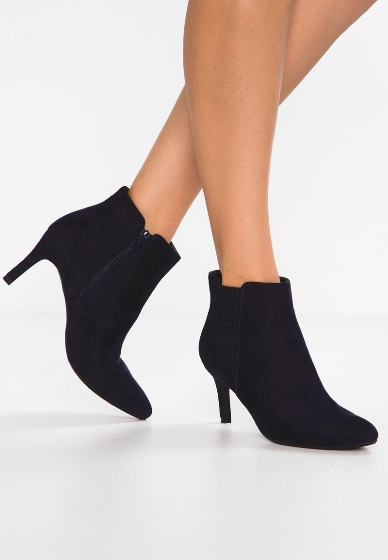 Femme portant des bottines en daim noir avec fermeture éclair latérale et talons fins de hauteur moyenne, debout sur un sol blanc.