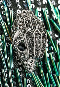 Decorazione a forma di mano in argento con cristalli neri e dettagli intricati, su uno sfondo nero testurizzato con paillettes iridescenti.