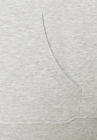 Felpa grigia chiara caratterizzata da un tessuto morbido e testurizzato, con una tasca frontale e orlo a coste. Sono visibili dettagli delle cuciture.