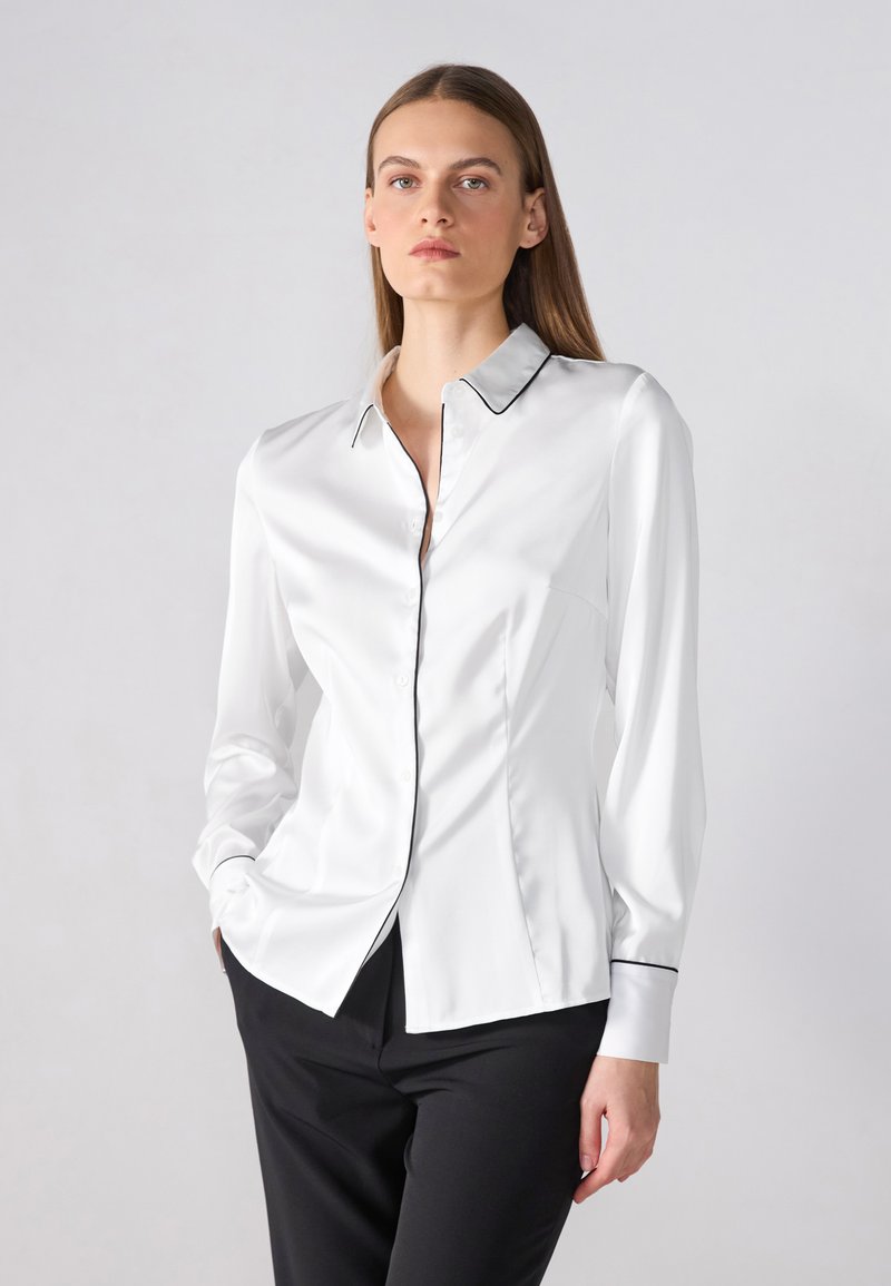 Camicia bianca a maniche lunghe realizzata in tessuto morbido, con colletto classico, bordi neri e design aderente con chiusura a bottoni.