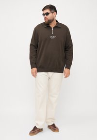 Sweatshirt zippé marron, logo blanc, associé à un pantalon beige clair et des baskets marron. Le modèle porte des lunettes de soleil, se tenant devant un fond uni.