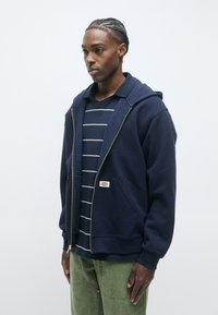 Felpa con zip di colore blu navy realizzata in tessuto morbido, con cappuccio, tasche frontali e una piccola etichetta con logo. Indossata sopra una maglietta a righe.
