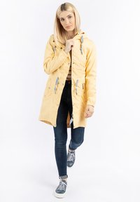 Jachetă galbenă impermeabilă cu glugă, închidere cu fermoar, detalii cu șnur și buzunare laterale, purtată cu blugi negri și pantofi sport.