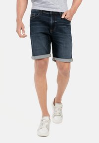 Mørkeblå denimshorts med opbrettede kanter, fem lommer og let falmet. Båret med hvide sneakers og en grå t-shirt.