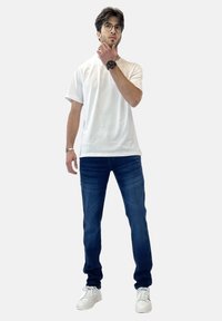 Camiseta blanca, jeans azules y zapatillas blancas. Persona de pie con la mano en la barbilla, usando gafas y un reloj oscuro. Fondo liso.