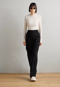Hvit rippet langermet topp med volangkrage og svarte skreddersydde bukser. Modellen har på seg mørke solbriller og hvite sko, og står på tregulv.