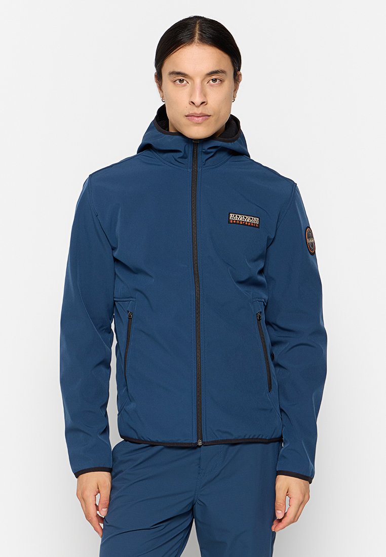 Hombre con chaqueta con capucha azul de cremallera, con bolsillos con cremallera y parches de logo en el pecho y la parte superior de la manga, de pie frente a un fondo liso.