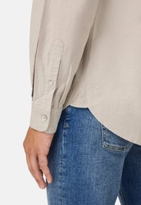 Camisa de manga larga beige con puños abotonados, que presenta un dobladillo redondeado y un detalle de logo cerca del dobladillo, combinada con jeans de mezclilla azul.