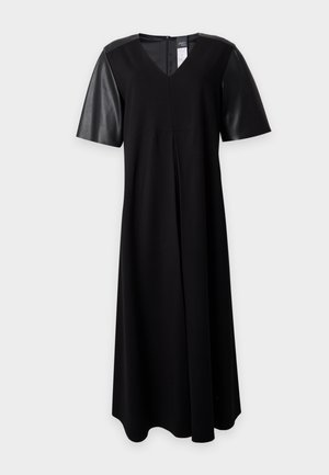 Femme debout portant une robe midi noire à manches courtes et des bottines noires, une main dans la poche, sur un fond clair uni.