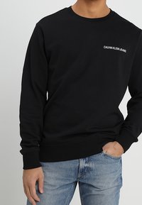 Man wearing a black Calvin Klein Jeans sweatshirt and light blue jeans, with one hand in his pocket against a plain background.