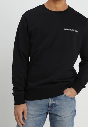 Man in een zwart Calvin Klein Jeans sweatshirt en lichtblauwe jeans, met één hand in zijn zak tegen een effen achtergrond.