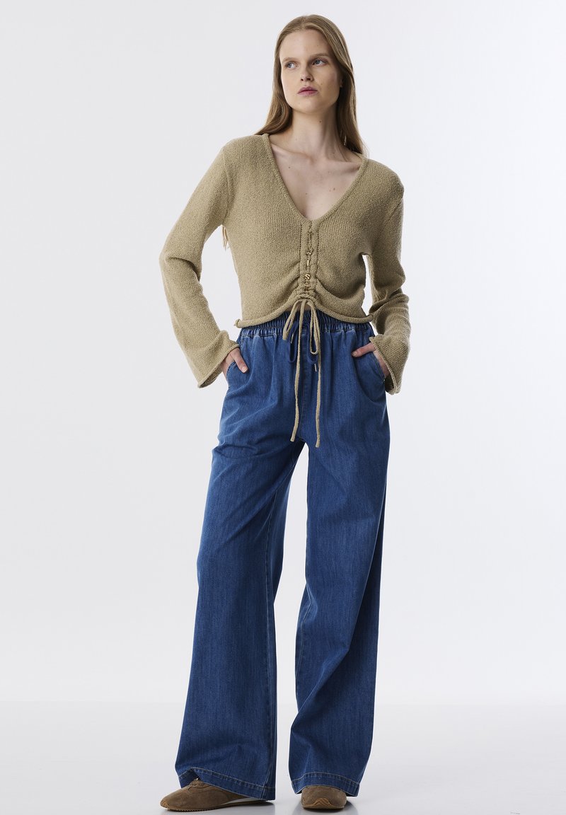 Beige gebreid top met een V-hals en een trekkoorddetail. Blauwe wijde denim broek met een elastische tailleband en zijzakken.