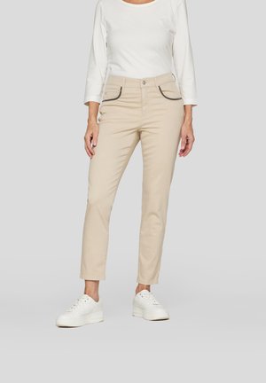 Beige Baumwollhose mit Seitentaschen, kontrastierenden Nähten, slim fit und schmal zulaufendem Bein; kombiniert mit einem weißen Langarmoberteil.