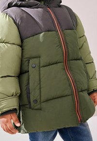 Chaqueta acolchada verde y negra con capucha, que presenta un zipper naranja, bolsillos laterales y tela texturizada para aislamiento.