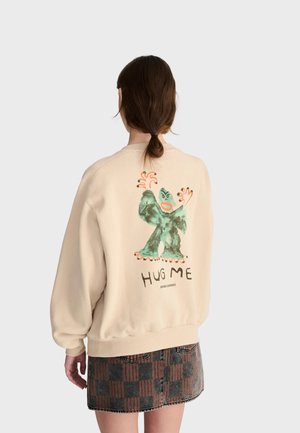 Person med brunt hår bundet i en lav hestehale iført en beige sweatshirt med en grøn tegneseriefigur og teksten "HUG ME" på ryggen.