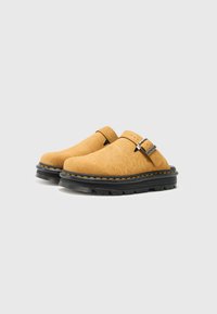 Bež slip-on klompe izrađene od teksturiranog materijala, s crnom gumenom potplatom koja ima narančastu šav i detaljom od srebrne kopče.