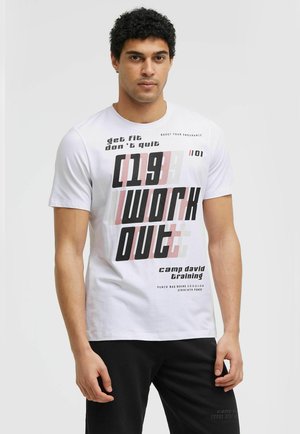 Mann mit lockigem Haar, der ein weißes T-Shirt mit schwarzem und rotem Workout-Schriftzug und schwarze Hose trägt, steht vor einem einfarbigen hellen Hintergrund.
