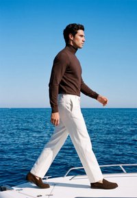 Homme en col roulé marron et pantalon blanc marchant avec assurance sur le pont d’un bateau avec une mer bleue calme et un ciel clair en arrière-plan.