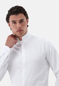 Camicia bianca con colletto a fascia, maniche lunghe e tessuto texturizzato. Presenta piccoli bottoni avorio e un design aderente.