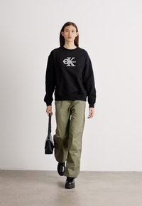 Čierna mikina s logom "Calvin Klein", olivovo zelené široké nohavice a čierne robustné čižmy. Model drží čiernu kabelku a kráča vpred.