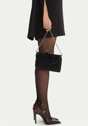 Žena v černých šatech, která nosí průhledné černé punčocháče a černé lodičky s špičatou špičkou, drží černé psaníčko s řetízkovým páskem a látkovou květinou.