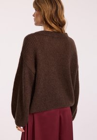 Brązowy sweter dzianinowy o luźnym kroju i prążkowanej teksturze, z szerokim dekoltem i długimi rękawami, w połączeniu z głęboko burgundową spódnicą.