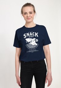 Marinblå t-shirt med en vit grafisk design som visar en haj och texten "SNACK ATTACK" ovanför detaljer från ett nautiskt klubb. Kort ärm och avslappnad passform.