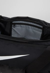 Czarna torba podróżna z dużym białym logo Nike, z główną komorą zapinaną na zamek oraz mniejszą wewnętrzną kieszenią z szarego materiału.