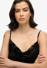 Camisole noir à motif floral avec des bretelles fines, présentant un décolleté drapé et une texture transparente. Le modèle a les bras croisés, mettant en valeur le design.