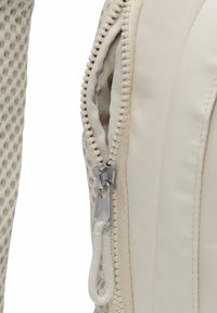 Beige tygväska med en texturerad, nätlika sida. Har en kontrasterande dragkedja med en silverfärgad dragflik, delvis öppen, vilket avslöjar insidan.