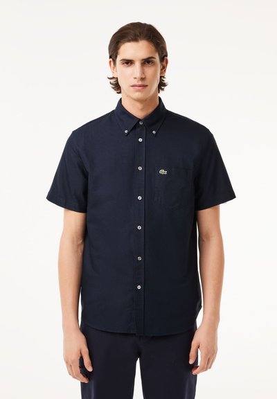 Chemise à manches courtes, de couleur bleu marine foncé, avec un col boutonné, une poche poitrine unique et le logo Lacoste près de la poche. Matériau en coton mélangé doux.