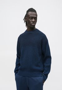 Tengerészkék kötött pulóver texturált mintával, kerek nyakkivágással és enyhén túlméretezett szabással, sötét nadrággal párosítva.