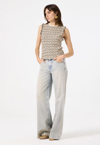 Stricktop mit schwarz-beigem Wellenmuster, mit Rüschenärmeln; hellblaue weit geschnittene Jeans. Das Modell posiert mit den Händen in den Taschen.