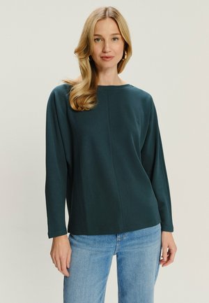 Kobieta z blond włosami, nosząca złote kolczyki w kształcie kół, ciemnozielony top z długim rękawem i jasnoniebieskie dżinsy, stojąca na jednolitym tle.