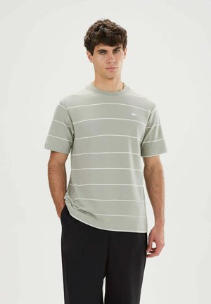 Jonge man met kort krullend haar, die een lichtgrijs gestreept T-shirt en zwarte broek draagt, staand met één hand in zijn zak.