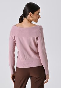 Maglione rosa scollato con collo a costine e maniche lunghe, caratterizzato da una vita aderente e una texture liscia. Abbinato a pantaloni marroni.
