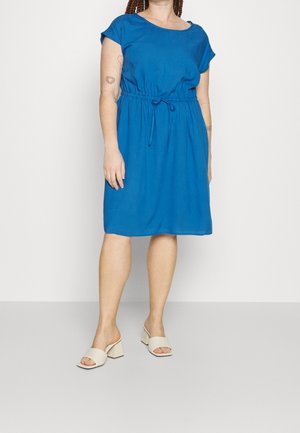 Kvinde iført en knælængde, kortærmet blå kjole med bindebånd i taljen, beige blokhælede åbne sandaler og synlige arm-tatoveringer.