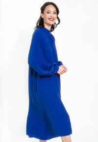 Langes blaues Kleid aus leichtem Stoff mit stehendem Kragen, Puffärmeln und Knopfgeschäften, das eine lockere, fließende Silhouette aufweist.