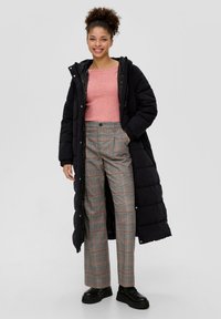 Gepolsterter schwarzer langer Mantel, hellrosa gerippter Pullover, graue karierte weit geschnittene Hose mit orangefarbenen Akzenten und schwarze Plateauschuhe.