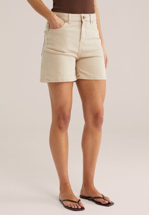 Žena oblečená v béžových džínových šortkách s vysokým pásom, hnedom topu a čiernych žabkách, stojaca s jednou rukou v vrecku na jednoduchom pozadí.