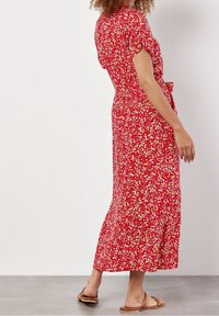 Femme en robe midi rouge à fleurs avec manches courtes et ceinture nouée, portant des sandales plates marron, debout sur un sol gris contre un mur blanc.