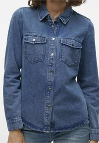 Kvinde iført en langærmet blå denimskjorte med knapper på brystlommerne og trykknapper foran, kombineret med blå jeans.