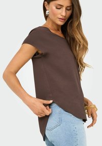 Donna che indossa una maglietta marrone a maniche corte con orlo asimmetrico e jeans azzurri, accessoriate con orecchini in argento e un bracciale traslucido.