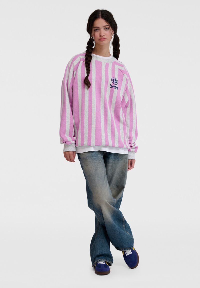 Överdimensionerad tröja i rosa och vita ränder med grå detaljer, bärs över en vit skjorta, kombinerad med ljusblå utsvängda jeans och lila sneakers.
