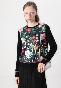 Colorful floral-patterned sweater with black sleeves and trim, featuring a rounded neckline and textured fabric. The model holds a small pink bag.