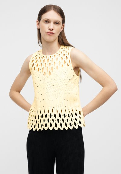 Jeune femme aux cheveux bruns raides portant un haut jaune sans manches avec des motifs découpés et un pantalon noir, posant avec les mains sur les hanches.