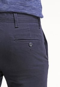 Detailní záběr na osobu, která má na sobě tmavě modré kalhoty s knoflíkovou zadní kapsou a modro-bíle pruhovanou košili zasunutou dovnitř.